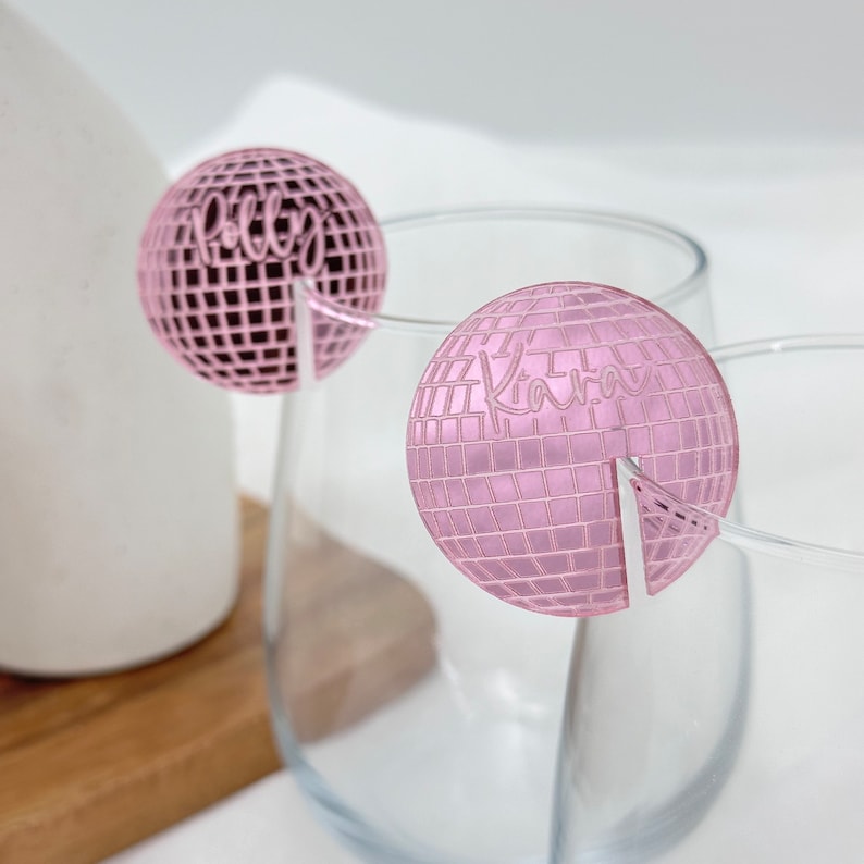 May include: Two pink disco ball-shaped drink charms with names, "Polly" and "Kara", etched on them. The charms are designed to sit on the rim of a glass. The background is a blurred white and light gray.