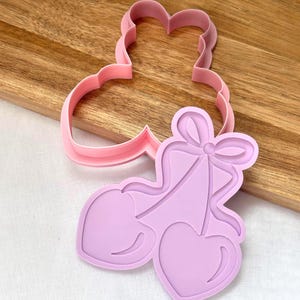 May include: Pink plastic cookie cutters. One is in the shape of a bell with two hearts and a bow, the other is a bell shape. The cutters are on a wooden surface.