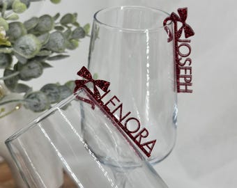 Puede incluir: Dos copas de vino sin tallo transparentes con etiquetas con lazo de purpurina roja que dicen "LENORA" y "JOSEPH". Las etiquetas están adheridas al borde de las copas. El fondo incluye vegetación.