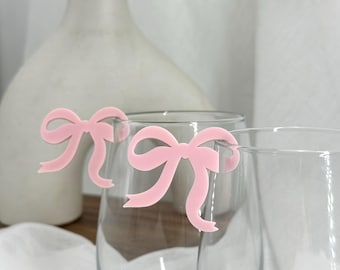 Puede incluir: Dos copas de vino transparentes con adornos rosas en forma de lazo. Los adornos están sujetos al borde de las copas. Un jarrón blanco está en el fondo, junto con un paño blanco.