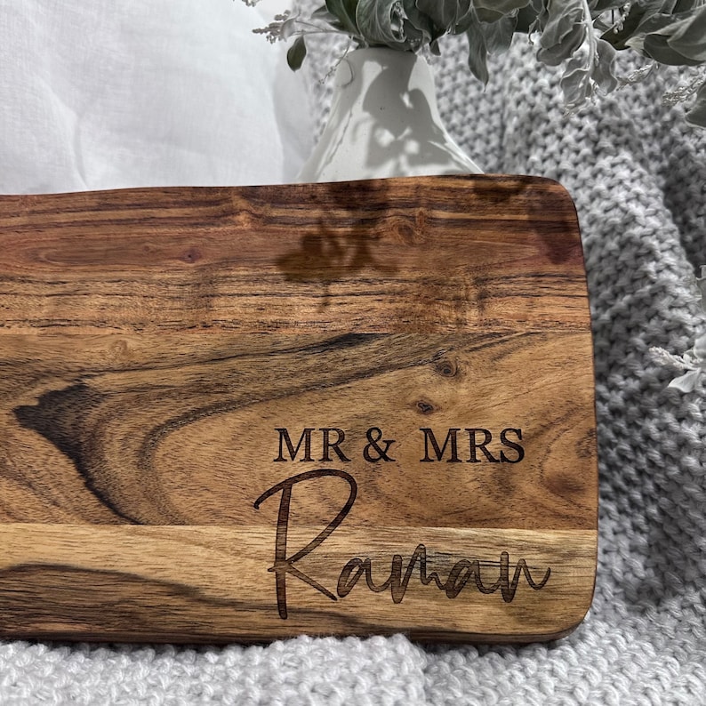 May include: A rectangular wooden cutting board with a natural wood grain finish. The words "MR & MRS" are engraved above the name "Raman" in a script font. The board is likely for serving or display.