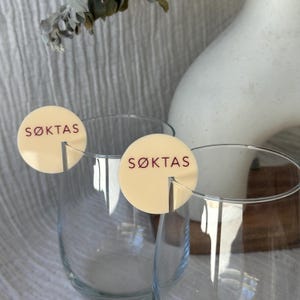 May include: Two clear wine glasses with cream-colored circular tags that read "S&Oslash;KTAS" in burgundy lettering. The tags are attached to the glasses. A white vase and dried foliage are in the background.