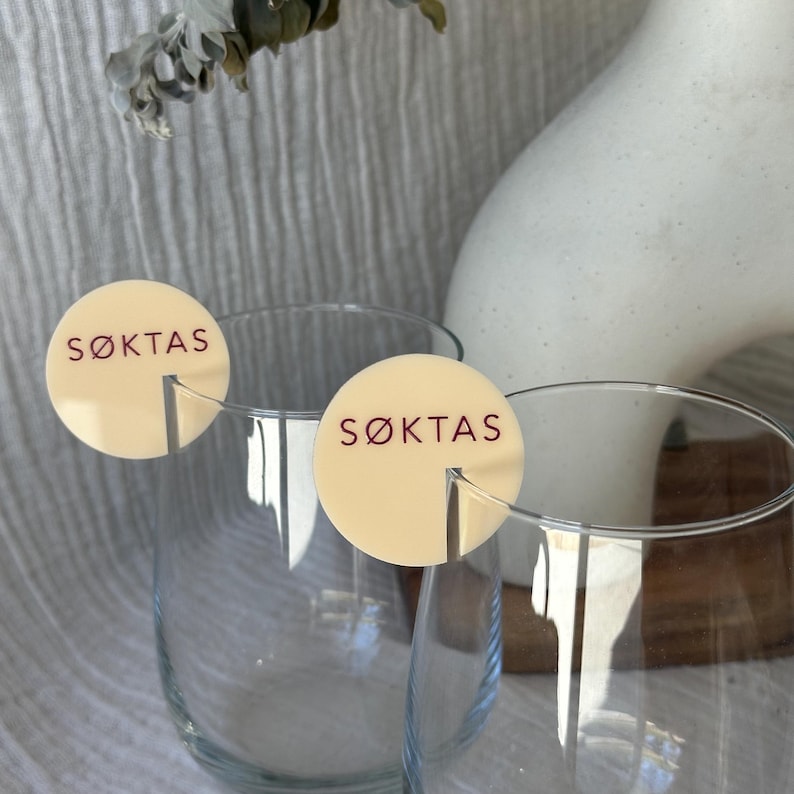 May include: Two clear wine glasses with cream-colored circular tags that read "S&Oslash;KTAS" in burgundy lettering. The tags are attached to the glasses. A white vase and dried foliage are in the background.