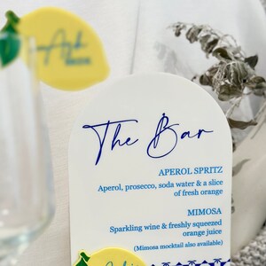 Może przedstawiać: Biały akrylowy szyld barowy z niebieskim napisem "The Bar". Na szyldzie wymienione są koktajle Aperol Spritz i Mimosa. Ż&oacute;łty mieszadełko do napoj&oacute;w w kształcie cytryny znajduje się na pierwszym planie.