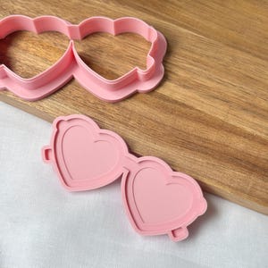 May include: Pink heart-shaped sunglasses cookie cutter and a matching outline cutter. The cutters are made of plastic and are on a wooden surface. The sunglasses have heart-shaped lenses and a decorative edge.