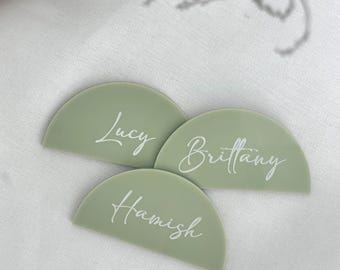 Könnte beinhalten: Drei halbkreisförmige Tischkarten in Salbeigrün mit weißen kursiven Namen: Lucy, Brittany und Hamish. Die Karten liegen auf einer weißen, strukturierten Oberfläche, ideal für Veranstaltungen.