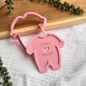 Puede incluir: Cortador de galletas rosa con forma de body con las palabras "FIRST EDITION" y un corazón. El cortador es de plástico y tiene un asa en forma de nube. El diseño es para galletas con temática de bebé.