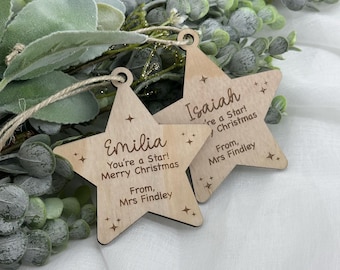 May include: Two wooden star-shaped Christmas decorations. One says "Emilia, You're a Star! Merry Christmas, From, Mrs Findley." The other says "Isaiah, 're a Star! Christmas, From, Mrs Findley." Each has a hole for hanging.