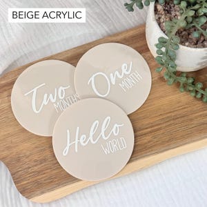 Peut inclure: Trois disques de jalons pour bébé en acrylique beige avec lettrage blanc. Les disques sont ronds et comportent les phrases "Hello World", "One Month" et "Two Months". Le texte "Beige Acrylic" est en haut à gauche.