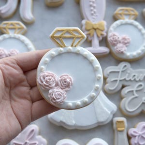 Peut inclure: Un biscuit décoré en forme de bague de fiançailles. La bague présente un diamant doré, une base blanche avec des perles et trois décorations de roses roses. D'autres biscuits sont visibles en arrière-plan.