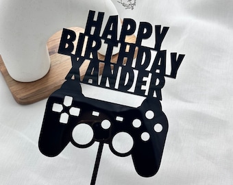 以下が含まれることがあります： 黒いアクリルケーキトッパー。 「HAPPY BIRTHDAY XANDER」の文字がコントローラーのデザインの上に配置されています。 このトッパーは誕生日ケーキ用にデザインされており、細い黒い棒で支えられています。