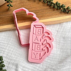 Puede incluir: Cortador de galletas de plástico rosa con forma de diseño de bloques de bebé. La palabra "BABY" está escrita en los bloques. El cortador tiene una parte superior en forma de corazón y un contorno rectangular.