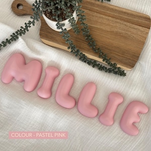 pink pastel-colored letters spelling out "MILLIE" on a white background, with a wooden cutting board and a potted plant in the background.