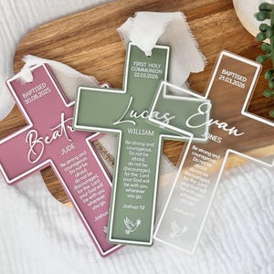 May include: Three cross-shaped ornaments in dusty rose, sage green, and clear acrylic. Each features white text including names, dates, and the Joshua 1:9 Bible verse. They are topped with white ribbons and display a small dove graphic at the base.