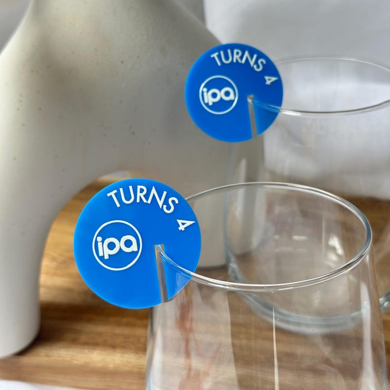 May include: Blue drink markers with the text "TURNS" and "IPA" in white. The markers are placed on the rim of a clear glass. A white ceramic vase is in the background.