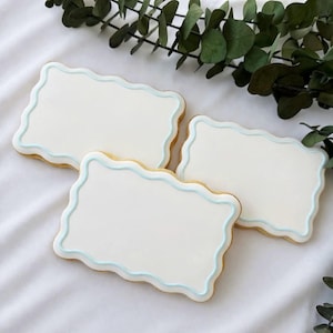 May include: Three rectangular sugar cookies with a scalloped edge, each frosted with white icing and a light blue border. The cookies are arranged on a white surface, with green foliage in the background.