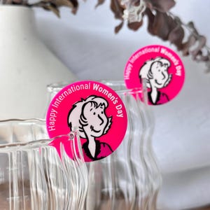 May include: Clear glass bottles with a wavy design, each adorned with a bright pink circular sticker. The stickers feature a cartoon illustration of a woman and the text "Happy International Women's Day" in white.