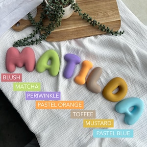 colorful letters spelling out the word "MATILDA" on a white cloth, with a potted plant and a wooden cutting board in the background.