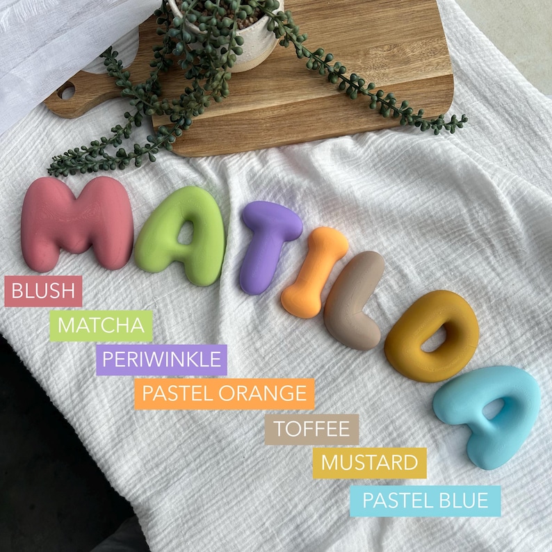 colorful letters spelling out the word "MATILDA" on a white cloth, with a potted plant and a wooden cutting board in the background.
