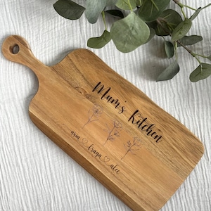 May include: Wooden cutting board with a handle, engraved with the words "Mum's Kitchen" and floral illustrations. The names "aria", "fraya", and "alec" are also engraved on the board. The board is made of light brown wood.