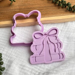 Puede incluir: Cortador de galletas de color lila claro con forma de pila de libros atados con un lazo. Un segundo cortador con forma de corazón está al lado del cortador de libros. Los cortadores son de plástico y están sobre una superficie blanca.
