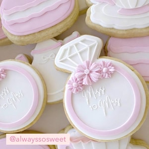 Op de afbeelding: Assortiment van versierde suikerkoekjes met een vrijgezellenfeestthema. Koekjes zijn onder meer een ringontwerp met roze bloemen en de tekst "Miss to Mrs", en koekjes in de vorm van een taart en een theepot. De koekjes zijn versierd met witte en roze glazuur.