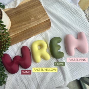 a wooden cutting board with the word "Wren" written on it, along with three different colored pastel pink, yellow, and olive letters. There are also some plants and a white cloth in the background.