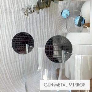 May include: Two clear wine glasses with gun metal mirror disco ball charms. The charms are round with a mirrored surface and are attached to the rim of the glasses. The text "GUN METAL MIRROR" is at the bottom of the image.
