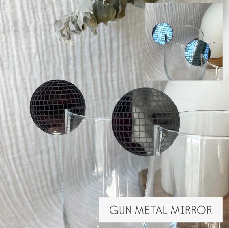 May include: Two clear wine glasses with gun metal mirror disco ball charms. The charms are round with a mirrored surface and are attached to the rim of the glasses. The text "GUN METAL MIRROR" is at the bottom of the image.