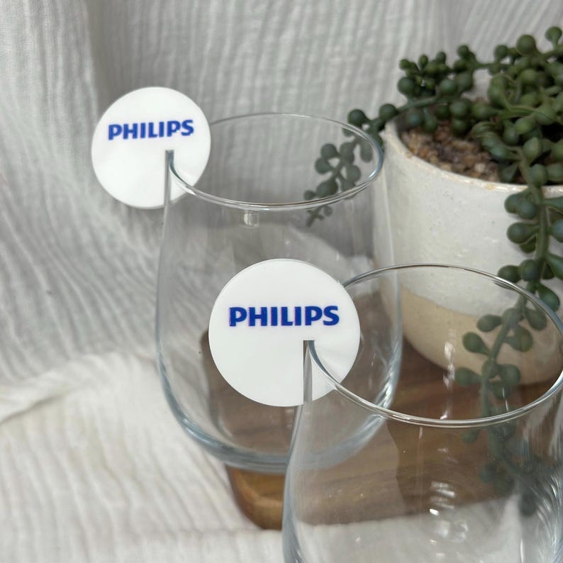 May include: Two clear wine glasses with white round coasters featuring the word "PHILIPS" in blue. The coasters are placed on the glasses. A small potted plant is visible in the background.