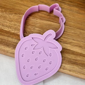 May include: A lilac strawberry-shaped cookie cutter. The cutter features a detailed strawberry outline with a stem and seed indentations. The cutter is made of plastic and is designed for baking and crafting.