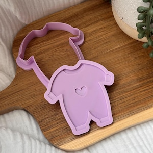 Puede incluir: Un cortador de galletas con forma de body de bebé de color lila claro con un corazón en el centro. El cortador es de plástico y tiene un asa. El cortador de galletas está sobre una tabla de cortar de madera.