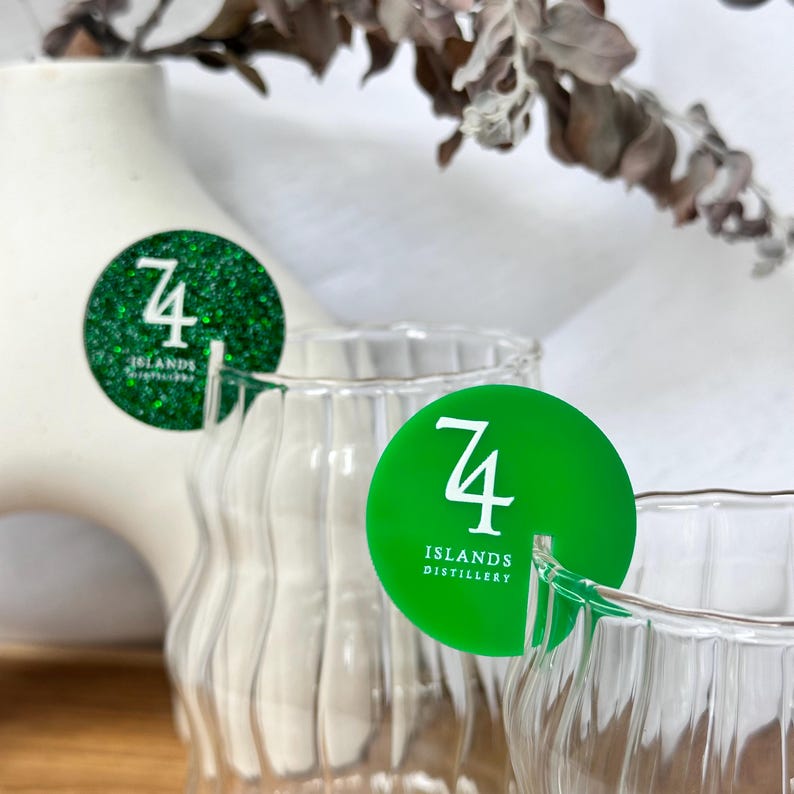 May include: Clear, ribbed glass with a green circular tag that reads "74 ISLANDS DISTILLERY" in white. One tag has green glitter. The background includes a white vase and dried foliage.