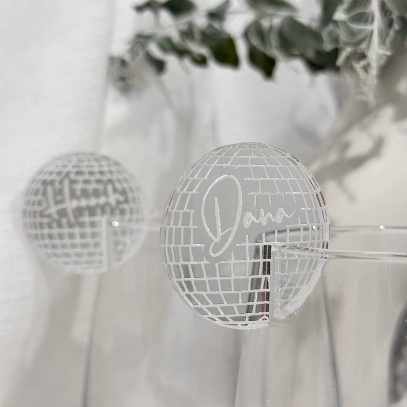 May include: Two clear acrylic disco ball-shaped place cards. The place card on the right has the name "Dana" etched in a cursive font. The disco balls are set against a white background with a hint of greenery.