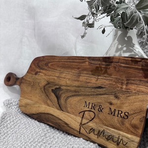 May include: A wooden serving board with a handle, featuring the engraved text "MR & MRS Raman". The board has a natural wood grain pattern and is set on a gray knitted surface, with a white vase and foliage in the background.