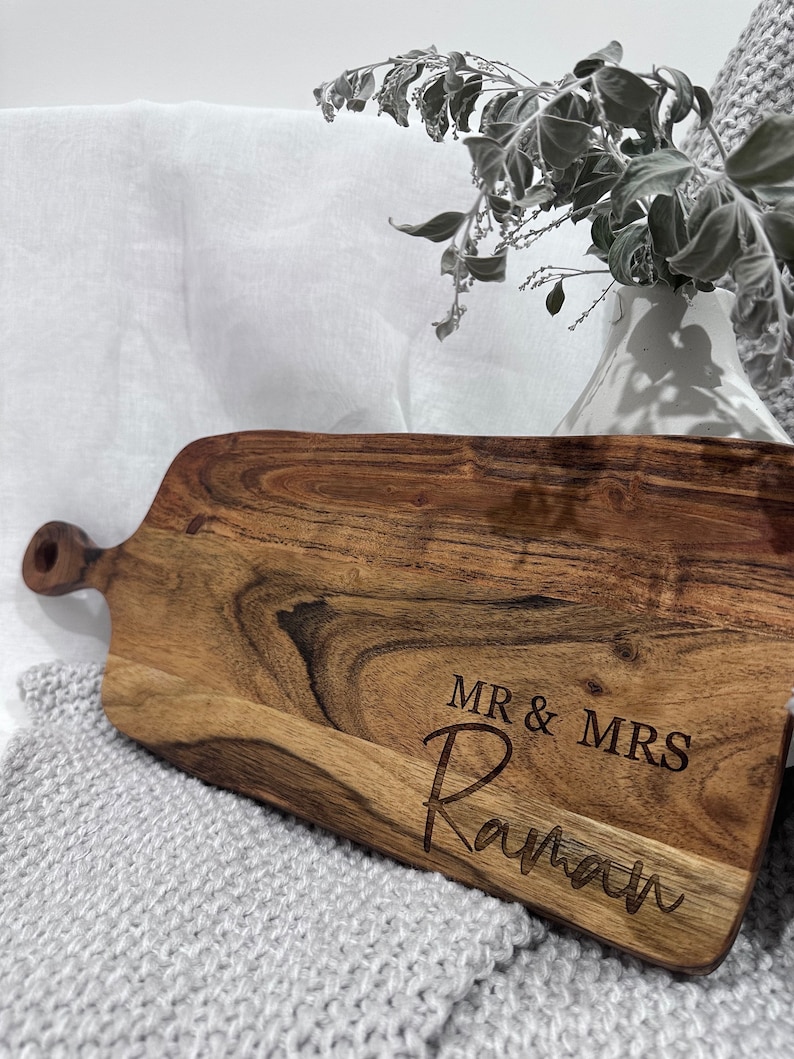 May include: A wooden serving board with a handle, featuring the engraved text "MR & MRS Raman". The board has a natural wood grain pattern and is set on a gray knitted surface, with a white vase and foliage in the background.