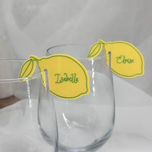 May include: Yellow lemon-shaped place cards with green accents and the names "Isabella" and "Eloise" in green script. The place cards are designed to hang on the rims of clear glasses.