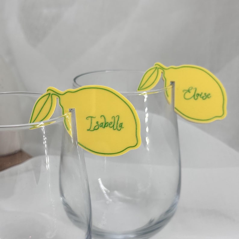 May include: Yellow lemon-shaped place cards with green accents and the names "Isabella" and "Eloise" in green script. The place cards are designed to hang on the rims of clear glasses.
