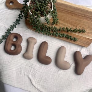 May include: Five individual letters spelling the name BILLY, crafted in smooth, rounded, matte-finish silicone in alternating shades of dark brown and light beige, arranged in a horizontal line on a white fabric surface.