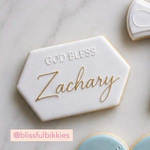May include: A hexagonal white iced biscuit featuring the embossed text GOD BLESS in white and the name Zachary written in elegant gold script. The biscuit has a smooth finish and is positioned alongside other partially visible decorated biscuits.