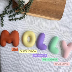 pastel-colored letters spelling out the name "Molly" on a white background, with a wooden board and some greenery in the background.
