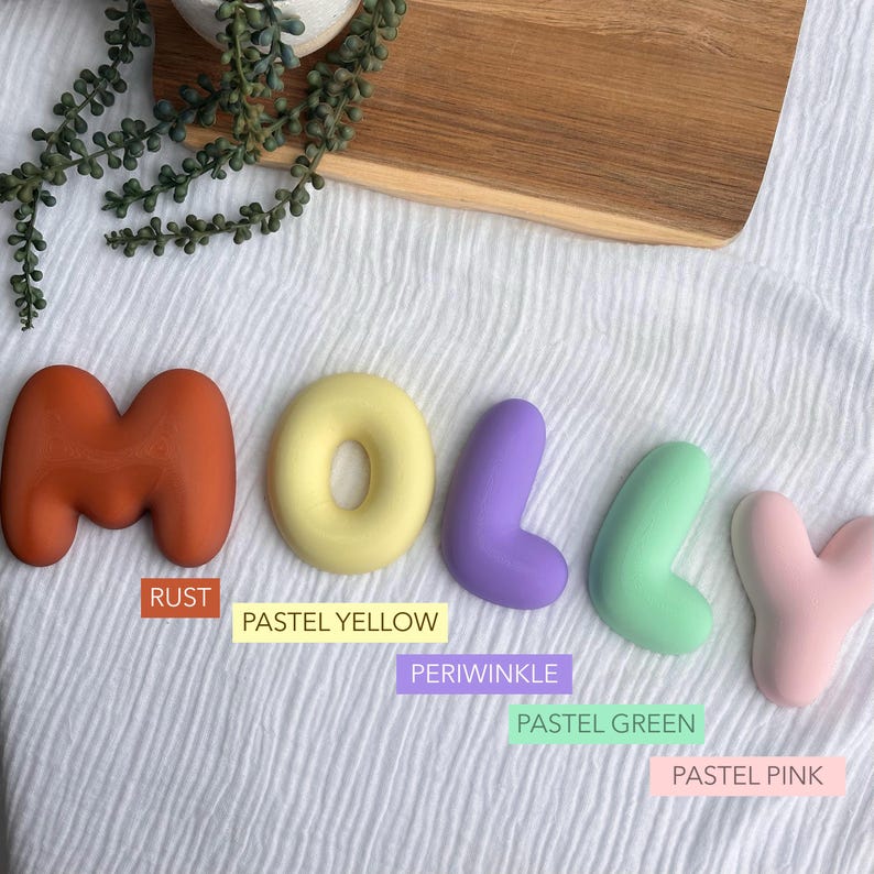 pastel-colored letters spelling out the name "Molly" on a white background, with a wooden board and some greenery in the background.