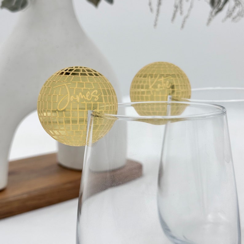 May include: Two gold disco ball-shaped drink charms with the names "James" and "Kate" etched on them. The charms are attached to the rims of clear wine glasses. A white vase and wooden tray are in the background.