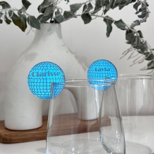 May include: Two clear wine glasses with blue disco ball-shaped drink charms. One charm says "Clarisse" and the other says "Layla". The charms have a grid pattern and are attached to the glasses with a silver wire.
