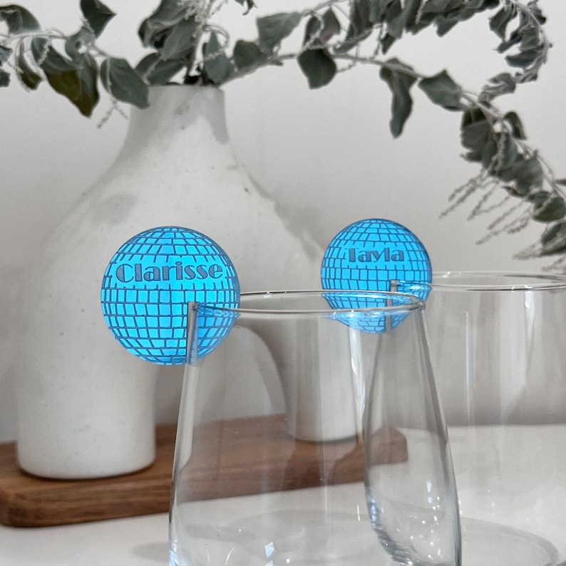 May include: Two clear wine glasses with blue disco ball-shaped drink charms. One charm says "Clarisse" and the other says "Layla". The charms have a grid pattern and are attached to the glasses with a silver wire.