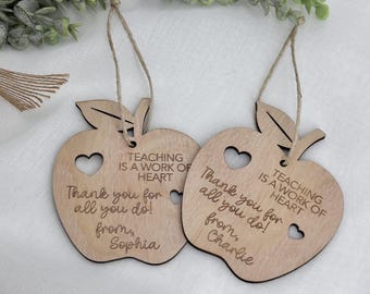 May include: Two wooden apple-shaped ornaments with heart cutouts and twine hangers. The ornaments are engraved with the phrases "Teaching is a work of heart" and "Thank you for all you do!" One is signed "Sophia" and the other "Charlie".