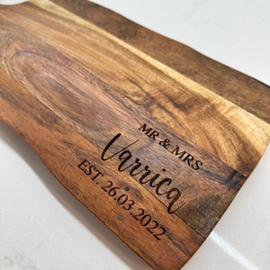 May include: A wooden cutting board with a natural edge. The board is engraved with the words "MR & MRS Varrica EST. 26.03.2022" in a stylish font. The wood grain is visible, showcasing a warm, brown color.