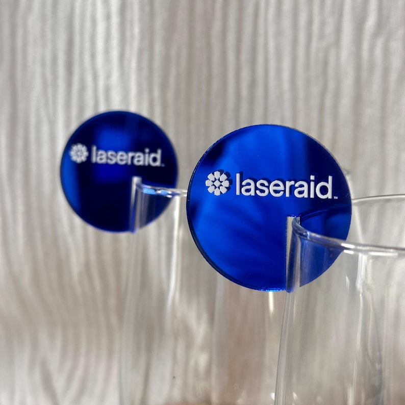 May include: Two blue circular drink markers with the text "laseraid" in white. The markers are attached to the rim of a clear glass. The background is a soft, white fabric.