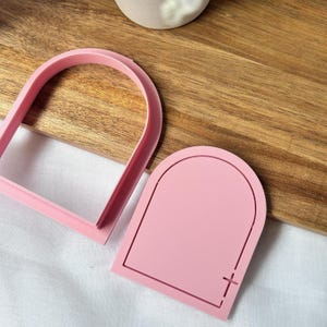 Puede incluir: Dos cortadores de galletas rosas con forma de ventana arqueada. Uno es un marco y el otro una forma sólida con un corte de cruz. Los cortadores están sobre una superficie de madera.