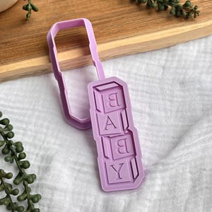 Puede incluir: Un cortador de galletas de color lavanda con forma de bloques apilados que deletrean "BABY". El cortador tiene un mango rectangular y está diseñado para cortar masa o fondant. La imagen está colocada sobre una tabla de cortar de madera y tela blanca.